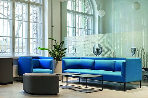 Ein offener, modern eingerichteter Raum am neuen Campus der Berlin School of Business and Innovation in Berlin Neukölln. Im Zentrum steht eine blaue Sitzgruppe von Kinnarps. Der Raum wird durch eine Glaswand unterteilt. Im Hintergrund sind die mehrere Meter hohen historischen Bogenfenster der alten Post zu erkennen.