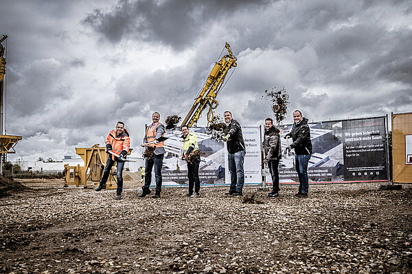 Sechs Personen in Arbeitskleidung auf der Baustelle des neuen NITRAS Logistikzentrums in Bedburg Mühlenerft. Jede Person hält einen Spaten in der Hand und schleudert Erde in Richtung der Kamera.