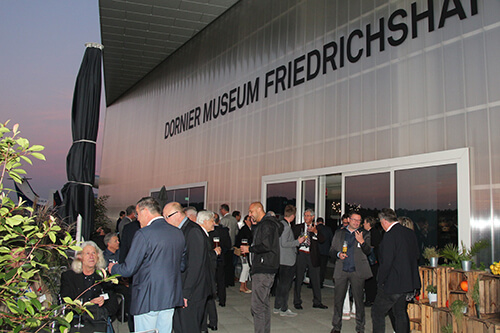 Außenansicht des Dornier Museums Friedrichshafen während der Feier zum 20-Jährigen Jubiläum des ECN 2021. Viele Menschen stehen vor dem Gebäude und unterhalten sich in kleineren Gruppen.