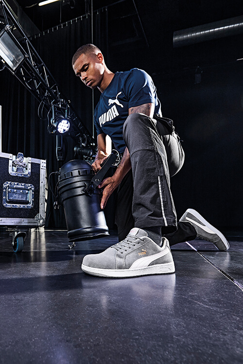 Ein Bühnentechniker mit Puma-Shirt und sportlichen Puma-Schuhen kniet auf dem Boden einer Bühne und hält einen Licht-Spot in der Hand.