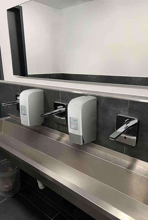 Ein langes Edelstahl-Waschbecken in einem Waschraum. Neben den Armaturen hängen zwei verschiedene Variomat-Touchless-Spender von PGP.