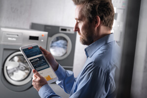 Ein Mann im Hemd steht vor einer Miele-Waschmaschine. Er blickt auf ein Tablet in seiner Hand, auf der das Portal "Miele MOVE" angezeigt wird.