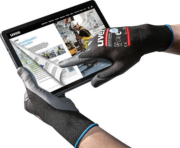 Nahansicht von zwei Händen, die Arbeitshandschuhe von UVEX tragen. Die linke Hand hält ein Tablet. Auf dem Bildschirm ist der digitale Katalog von UVEX abgebildet. Die rechte Hand blättert mit dem Zeigefinger auf dem Tablet zur vorherigen Seite.