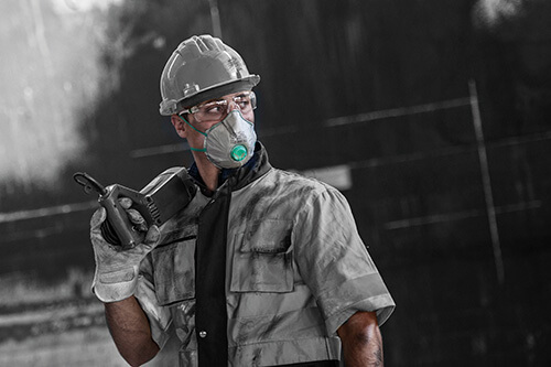 Eine Person in grauer Arbeitskleidung mit Schutzbrille und Bauhelm trägt eine graue FFP3-Maske von BLS.